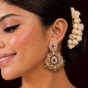 A close-up of a woman wearing an ornate gold-toned chandelier earring with red and green stones, against a dark brown background.