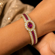 A close-up of a gold bangle bracelet with a central oval ruby gemstone and rows of round and baguette-cut rubies, worn on a person's wrist.