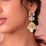 A close-up of a woman wearing a large gold-toned traditional jhumka earring adorned with a pink stone and white stones, against a soft pink background.