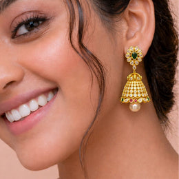 A close-up of a woman wearing a gold-toned traditional Indian jhumka earring with green, red, and white stones, and a pearl drop, shown on her ear.