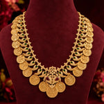 An ornate gold-plated traditional Indian coin necklace with green and red stone accents, displayed on a dark red velvet jewelry bust.