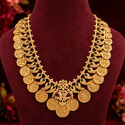 An ornate gold-plated traditional Indian coin necklace with green and red stone accents, displayed on a dark red velvet jewelry bust.