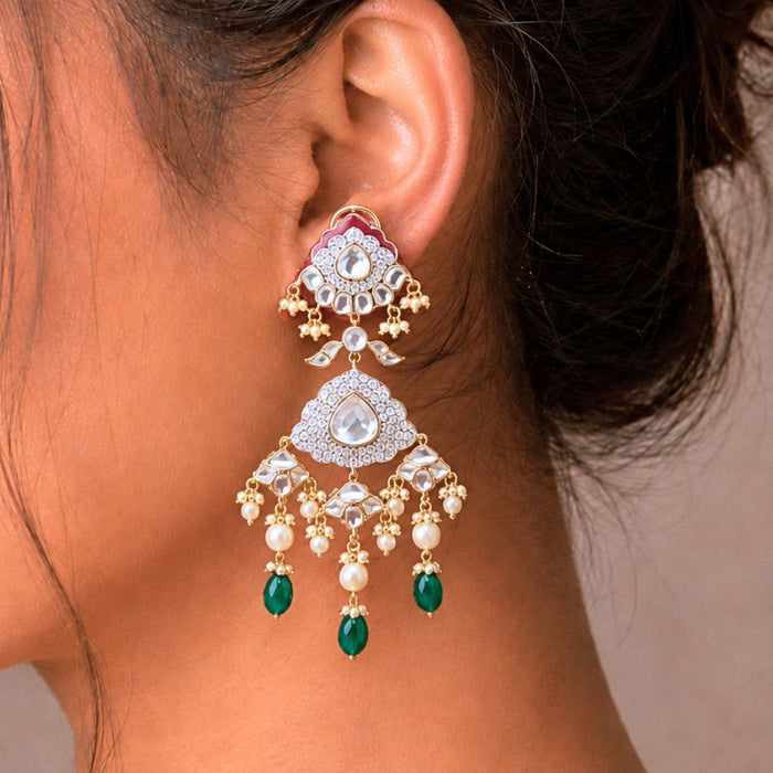 A close-up of a woman's ear wearing a large, ornate gold-toned Indian style dangle earring decorated with clear stones, white pearls, and green teardrop beads.