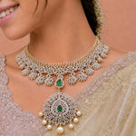 A close-up of a woman wearing an ornate gold and white stone choker necklace with a large matching pendant featuring green emerald accents and pearl-like drops, paired with a beige embroidered saree.