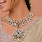 A close-up of a woman wearing an ornate gold and white stone choker necklace with a large matching pendant featuring green emerald accents and pearl-like drops, paired with a beige embroidered saree.
