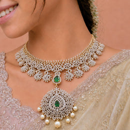 A close-up of a woman wearing an ornate gold and white stone choker necklace with a large matching pendant featuring green emerald accents and pearl-like drops, paired with a beige embroidered saree.